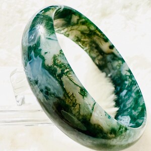May include: A translucent, moss agate bangle bracelet. The bracelet features a blend of green, white, and blue hues, with intricate patterns resembling natural landscapes. The polished surface reflects light, highlighting the stone's unique veining.