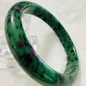 May include: A green jade bangle with dark green and red inclusions. The bangle is polished and has a smooth surface.