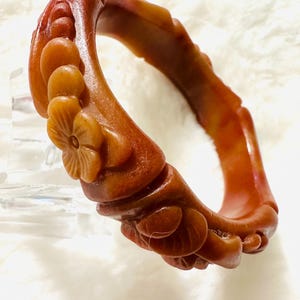 May include: A reddish-brown bangle bracelet featuring a carved floral motif with raised petals and circular accents. The surface has a smooth, semi-translucent finish with variations in color depth throughout the circular band structure.