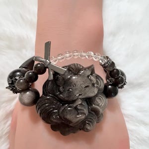 May include: A bracelet featuring a large, carved dark gray obsidian fox pendant with a metallic bow accent. The band consists of smooth, round dark gray obsidian beads, smaller textured dark metal beads, and a section of clear, round glass beads.