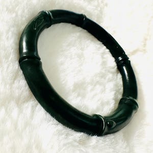 May include: Dark green bamboo-style bracelet. The bracelet is a solid, circular design with a glossy finish. The bamboo-style segments are connected by rounded, slightly raised sections. The bracelet is set against a white, fluffy background.