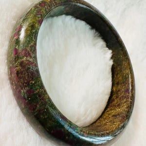 May include: A gemstone bangle bracelet. The bracelet is a mix of green, gold, and burgundy colors. The bangle is oval-shaped and appears to be made of a polished stone.