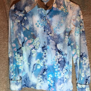 May include: A blue and white long-sleeved button-up shirt with a floral print. The shirt has a pointed collar and a single button closure. The fabric is soft and silky.