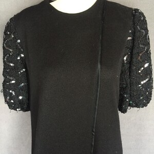 May include: Black short-sleeved sweater with sequined sleeves. The sleeves are adorned with black sequins and beads, creating a shimmering effect. The sweater has a round neckline and a vertical satin detail.