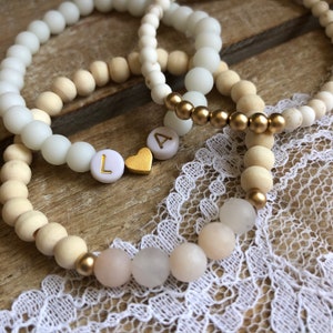 May include: Three beaded bracelets with white, light wood, and gold beads. One bracelet spells "LA" with gold letters and a gold heart charm. The bracelets rest on a wooden surface with a white lace trim.