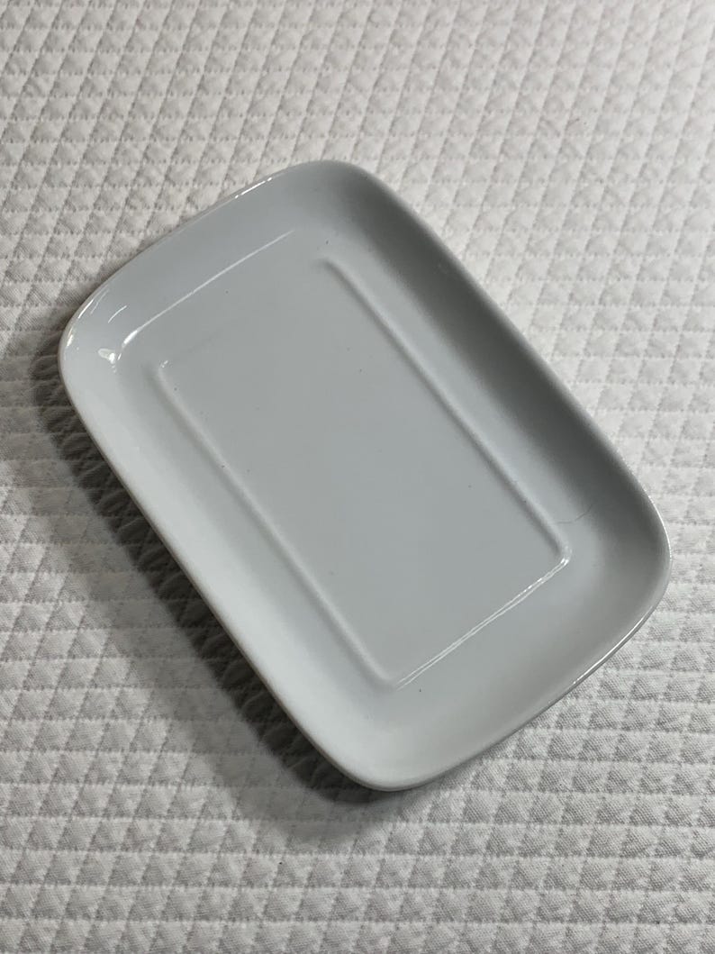 May include: A rectangular, pale grey ceramic dish with rounded corners. The dish has a slightly raised rim and a recessed centre. The surface is smooth and glossy. The dish is set against a white, textured background.