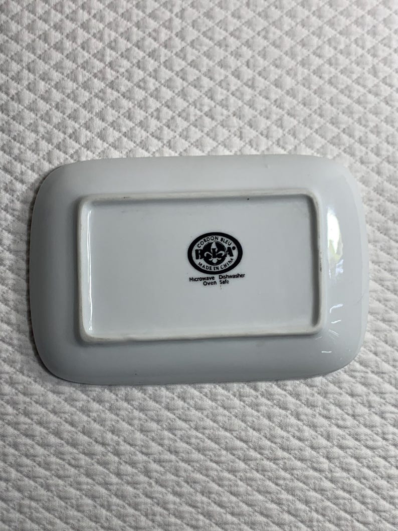 May include: White rectangular dish with rounded corners. The base features a black circular logo with text: "CORDON BLEU MADE IN CHINA Microwave Dishwasher Oven Safe."