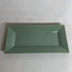 K&ouml;nnte beinhalten: Ein rechteckiger Servierteller aus salbeigr&uuml;nem Keramik. Der Teller hat eine gl&auml;nzende Oberfl&auml;che und einen leicht erh&ouml;hten Rand. Das Design ist schlicht und modern, geeignet zum Servieren von Vorspeisen oder kleinen Gerichten. Ma&szlig;e: ca. 30 cm lang und 15 cm breit.