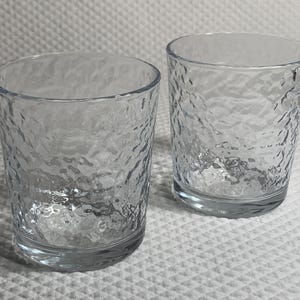 May include: Two clear, textured drinking glasses. The glasses have a unique hammered glass design. The glasses are cylindrical with a thick base. The glasses are empty and sitting on a white surface.
