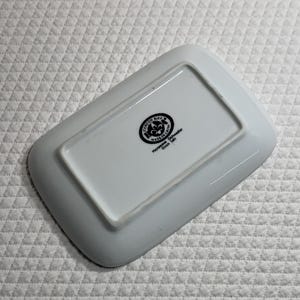 May include: A white rectangular ceramic dish with rounded corners. The underside features a black and white circular emblem with text, including "Cordon Bleu" and "Made in China". The dish is designed for microwave and dishwasher use.