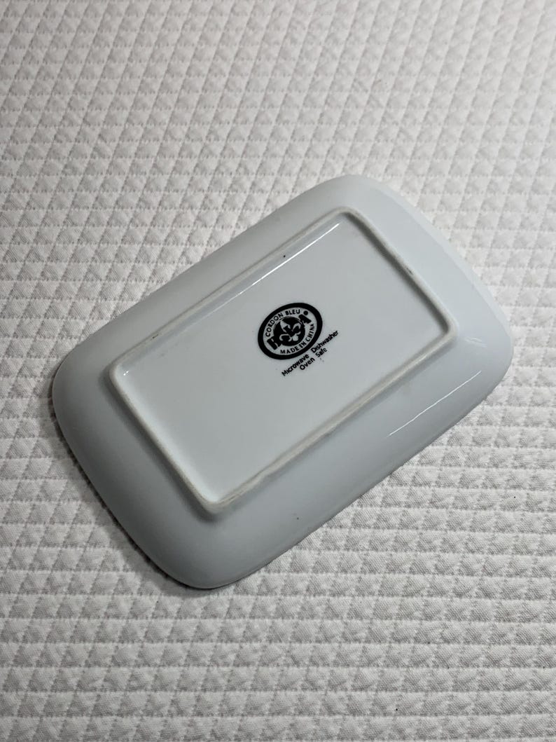 May include: A white rectangular ceramic dish with rounded corners. The underside features a black and white circular emblem with text, including "Cordon Bleu" and "Made in China". The dish is designed for microwave and dishwasher use.