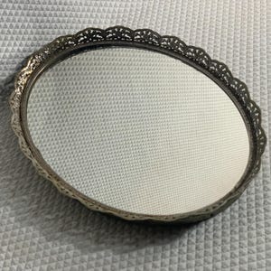 May include: An oval-shaped vanity tray featuring a reflective mirrored base and a decorative silver-toned metal rim with an intricate, openwork filigree pattern along the edge. The tray has a polished finish and a classic, symmetrical design.