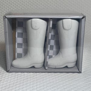 May include: A pair of white ceramic cowboy boot figurines displayed in a clear plastic box. The boots have a classic design with a rounded toe and a mid-height shaft. The box has a checkered pattern background.