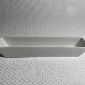 May include: A rectangular, light gray ceramic serving dish with slightly angled sides. The dish has a simple, modern design, perfect for serving appetizers or small dishes. The dish is set against a white textured background.
