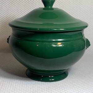 May include: A glossy, emerald green ceramic lidded serving bowl. The bowl has a rounded body, a pedestal base, and two small decorative handles. The lid has a rounded top with a small knob. The bowl is designed for serving food.