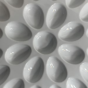 May include: Close-up of a white, glossy, ceramic serving tray with a raised, pebble-like design. The tray features a central circular pattern surrounded by oval-shaped indentations, creating a textured surface. The design is repeated across the tray.