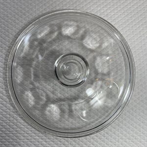May include: A clear, circular glass lid with a central knob. The lid has a raised rim and a series of subtle, decorative indentations. This kitchenware item is designed to fit on a pot or pan.