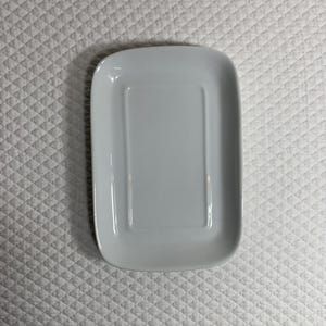 May include: A rectangular, light grey ceramic soap dish. The dish has a slightly raised rim and a recessed area in the centre. The overall shape is a rounded rectangle. The dish is clean and simple.