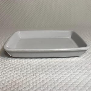 May include: A rectangular, light gray ceramic tray with slightly rounded corners. The tray has a smooth, glossy surface and is set against a white, textured background. The tray is designed for serving or display.