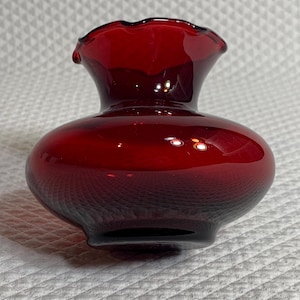 May include: A small, deep red glass vase with a rounded, bulbous body and a wide, ruffled rim. The glass is translucent and smooth, featuring a glossy finish that reflects light across its curved surface, creating a vibrant, rich crimson color.