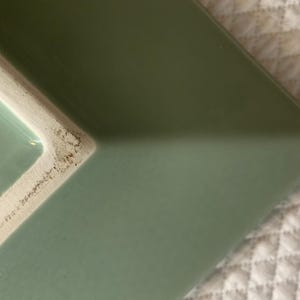 K&ouml;nnte beinhalten: Nahaufnahme eines rechteckigen Keramiktellers in Salbeigr&uuml;n. Der Teller hat eine leicht abgerundete Kante und einen wei&szlig;en Rand. Der Teller liegt auf einer wei&szlig;en, gesteppten Oberfl&auml;che.