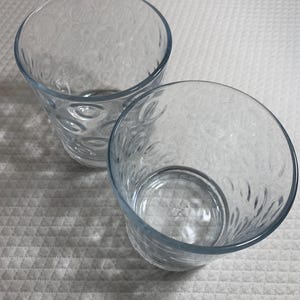 May include: Two clear, round drinking glasses with a textured pattern. The glasses are made of clear glass and have a slightly blue tint around the rim. The glasses are sitting on a white, textured surface.