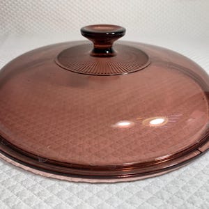 May include: A clear, round, glass lid with a dark reddish-brown hue. The lid features a small, rounded handle and a textured pattern on the top. The rim of the lid is also a darker shade of the same color. This kitchenware item is designed to fit on a pot or pan.
