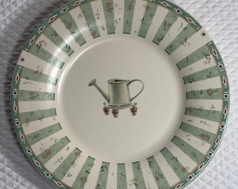 May include: A round ceramic plate with a cream-colored center featuring a watering can illustration. The plate has a green and white striped border with floral accents and a decorative rim. A charming addition to any kitchen.
