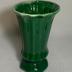 May include: A glossy, emerald green vase with a fluted design. The vase has a flared top and a pedestal base. The rim has a textured, off-white edge. The vase is approximately 8 inches tall.
