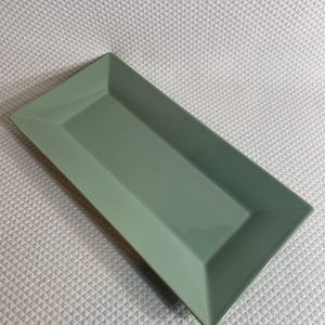 K&ouml;nnte beinhalten: Ein rechteckiger, salbeigr&uuml;ner Servierteller mit leicht erh&ouml;htem Rand. Der Teller hat ein schlichtes, modernes Design und besteht aus gl&auml;nzendem Material. Die Form ist ideal zum Servieren von Vorspeisen oder Desserts.
