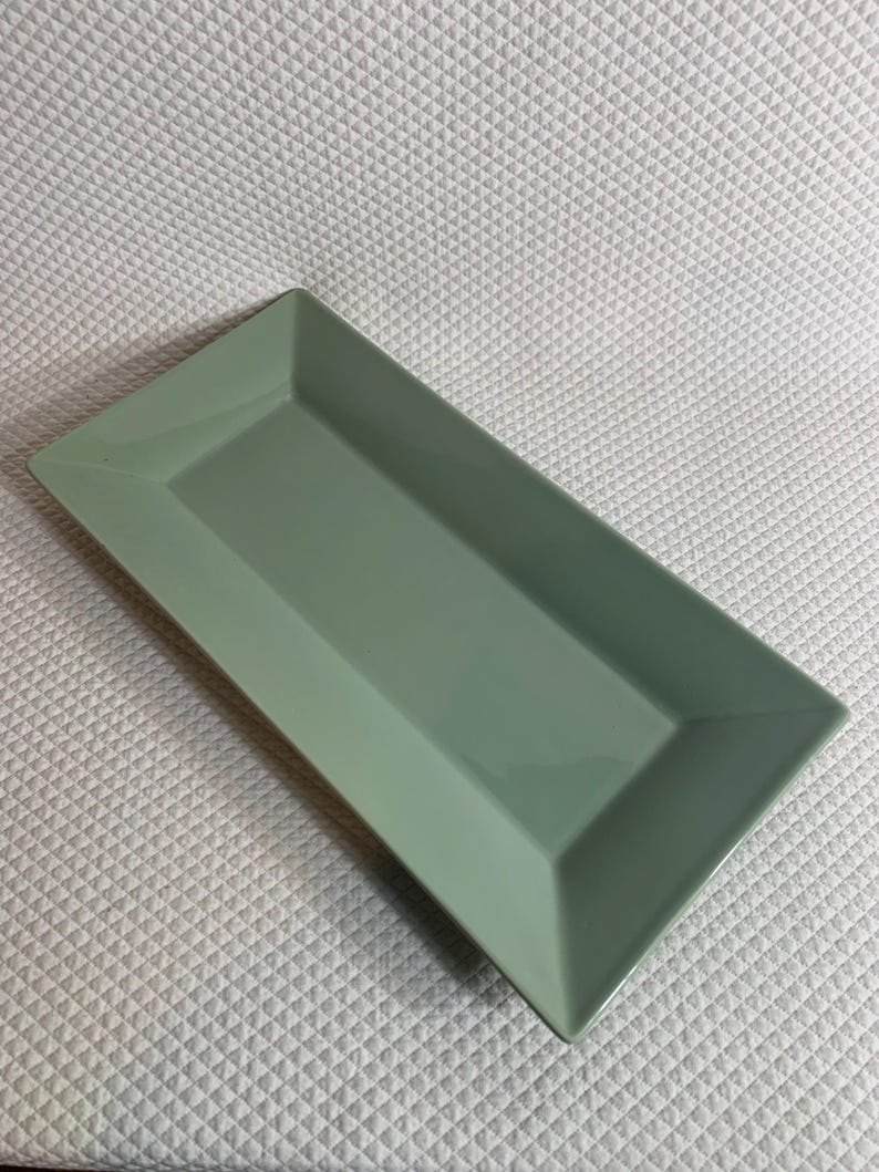 K&ouml;nnte beinhalten: Ein rechteckiger, salbeigr&uuml;ner Servierteller mit leicht erh&ouml;htem Rand. Der Teller hat ein schlichtes, modernes Design und besteht aus gl&auml;nzendem Material. Die Form ist ideal zum Servieren von Vorspeisen oder Desserts.