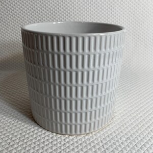 May include: A cylindrical ceramic planter with a glossy light gray finish. The exterior surface features a repeating vertical grid pattern consisting of raised rectangular bars arranged in columns around the entire circumference of the vessel.