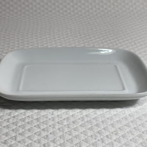 May include: A rectangular, white ceramic serving dish with rounded corners. The dish has a slightly raised edge and a smooth, glossy surface. The shape is ideal for serving appetizers or small portions.