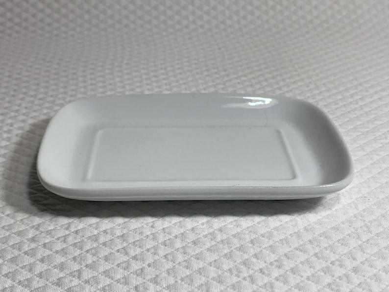 May include: A rectangular, white ceramic serving dish with rounded corners. The dish has a slightly raised edge and a smooth, glossy surface. The shape is ideal for serving appetizers or small portions.