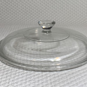May include: A clear glass lid with a small, rounded knob. The lid is circular with a slightly domed shape and a series of concentric rings around the edge. The knob is also clear and rounded. This kitchenware item is designed to fit on a pot or pan.