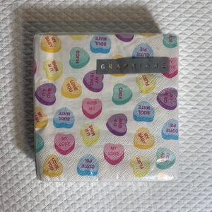 May include: A package of paper napkins with a repeating pattern of colorful candy hearts. Each heart has a different message, such as "Hugs," "Kiss Me," and "My Baby." The brand name "Graphique" is printed on a gray rectangle.