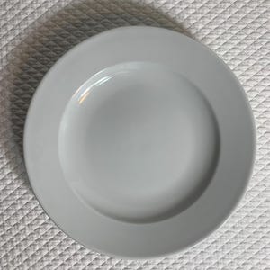 May include: A round, white ceramic plate with a wide rim. The plate is centered on a white, textured surface. The plate is a simple, elegant design, suitable for serving meals.