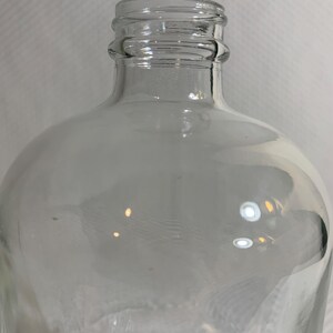 May include: A clear glass bottle featuring a rounded body and a narrow neck with a threaded top designed for a screw-on cap. The transparent glass construction allows for full visibility through the container, showcasing its smooth, curved shape.