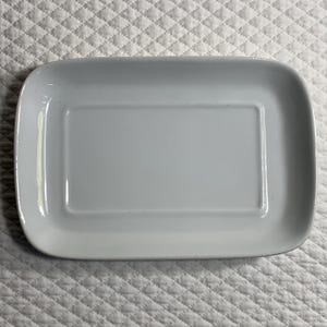May include: A rectangular, pale grey ceramic serving dish with rounded corners. The dish has a slightly raised inner rectangle. The background is a white, quilted fabric.