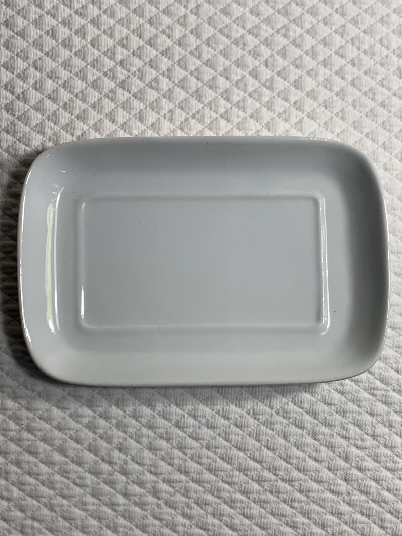 May include: A rectangular, pale grey ceramic serving dish with rounded corners. The dish has a slightly raised inner rectangle. The background is a white, quilted fabric.