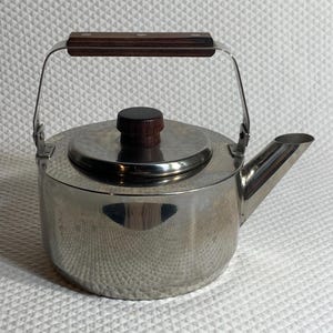 May include: A stainless steel tea kettle with a cylindrical body, a short angled spout, and a flat lid featuring a dark brown wooden knob. The kettle includes a pivoting metal handle with a dark brown wooden grip secured by three rivets.