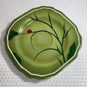 May include: A green ceramic plate with a leaf-like shape and a white rim. The plate features a painted design of a ladybug on a branch and green leaves. The plate has a diameter of approximately 8 inches.