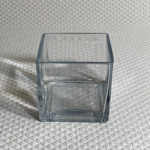 May include: A clear, square glass vase with a slightly curved base. The vase has a simple, modern design and is suitable for displaying flowers or other decorative items. The vase is empty and sits on a white, textured surface.