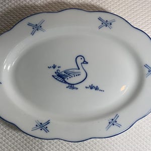 May include: White oval serving platter with a blue duck illustration and a scalloped edge. The plate has a blue border and decorative blue cross-like designs around the rim. A charming addition to any table setting.