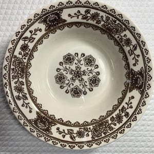 May include: A white ceramic bowl with a brown floral pattern. The bowl has a wide rim with a repeating floral and vine design. The center of the bowl features a cluster of flowers. The bowl is likely vintage or antique.