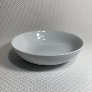 May include: A simple, round, light gray ceramic bowl. The bowl has a wide, shallow design and a smooth, glossy finish. The bowl is suitable for serving soups, salads, or other dishes. The bowl is sitting on a white, textured surface.