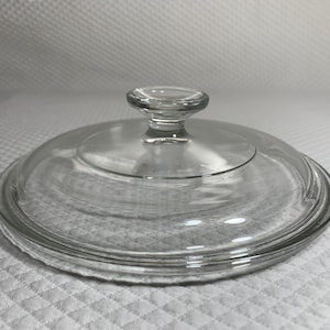 May include: A clear glass lid with a round shape and a small, rounded handle. The lid has a slightly raised edge and is designed to fit on a pot or pan. The glass is transparent, allowing visibility of the contents.