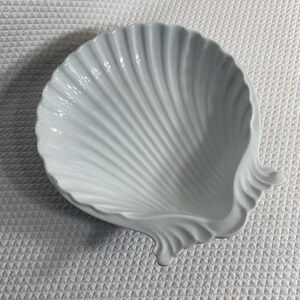 May include: A white ceramic dish shaped like a scallop shell. The bowl features a glossy finish with deep, radiating ridges that extend from the hinge area to the scalloped outer edge, creating a textured, fan-like appearance for the vessel.