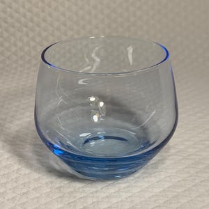 May include: A clear, stemless glass with a rounded bowl and a vibrant blue hue. The glass has a smooth, polished surface and a thick base. The rim of the glass is also blue. The glass is designed for drinking.