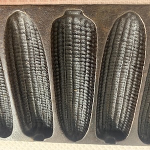 May include: A cast iron corn bread mold featuring five distinct, elongated, ear-of-corn shaped cavities. The metal surface is dark, textured, and matte, with each mold section showing detailed kernel patterns arranged in vertical rows.